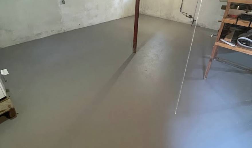 Basement-Epoxy-Floor A basement with a freshly painted gray concrete floor showcases recent basement concrete resurfacing, complemented by white walls, a red support pole, and part of a wooden shelving unit visible on the right.