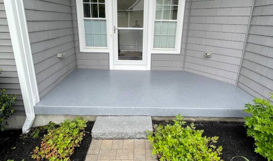 Concrete-Resurfacing-Patio A gray concrete porch, recently enhanced with patio concrete resurfacing, features a stone step leading to a white door with glass panels, surrounded by gray siding and small green shrubs.