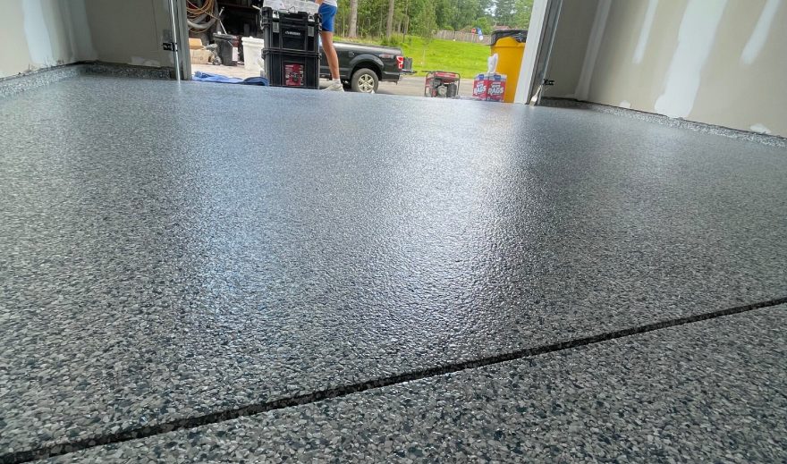A newly finished epoxy-coated garage floor with a speckled gray surface showcases expert Garage Concrete Resurfacing, inside a garage with drywall and an open door leading outside.