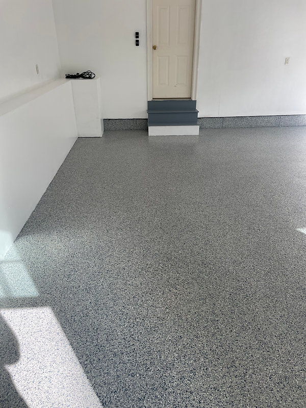 An empty garage with a clean, speckled epoxy floor from recent concrete resurfacing, white walls, a closed beige door with blue steps, and some cables on a white ledge.