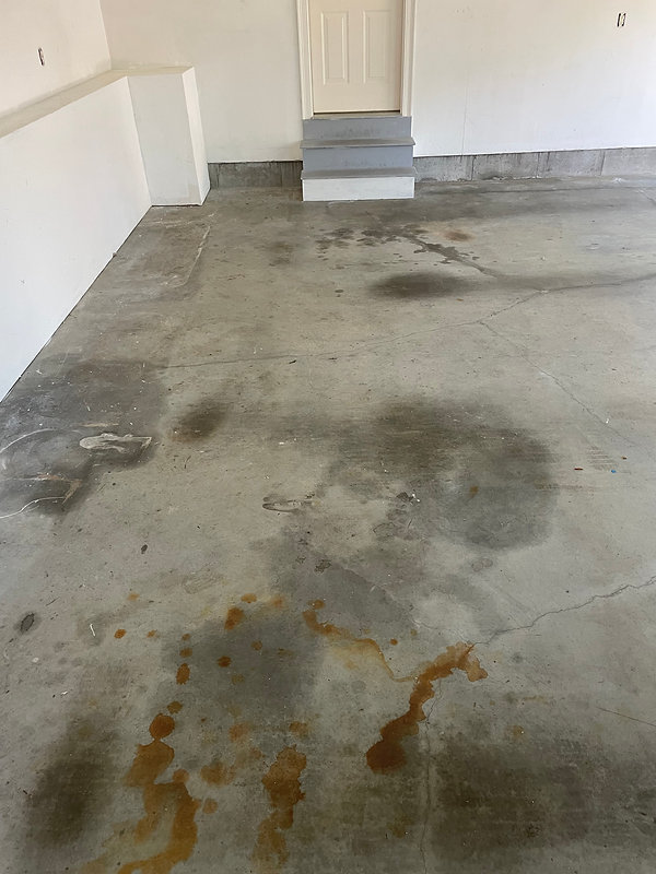 A garage with a concrete floor showing dark stains and patches, indicating the need for concrete resurfacing, leading up to a white door with three steps.