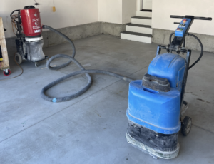 A garage set up for concrete resurfacing, featuring concrete grinding equipment—a blue floor grinder and a red vacuum machine connected by hoses. Shelving and a blue tarp are visible above.