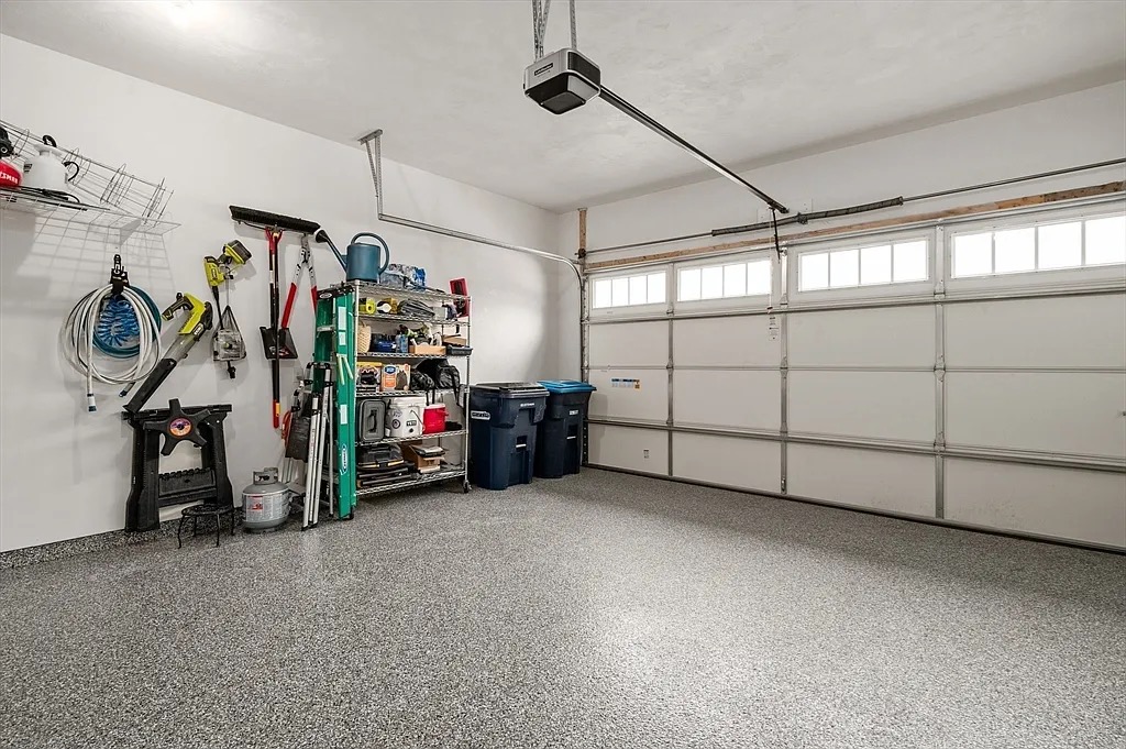 A clean, organized garage with concrete resurfacing, wall shelves, yard tools, storage bins, and two large trash cans next to a closed double garage door.