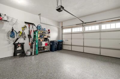 Neatly organized garage with shelves holding tools and storage bins, cleaning supplies on the wall, a closed double garage door, and a smooth surface that shows how epoxy flooring works to create a clean, durable finish.